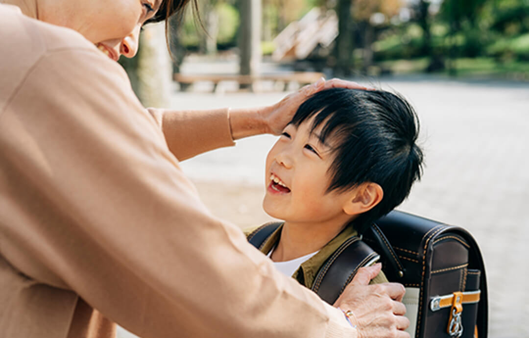 ランドセルを背負った小学生くらいの男の子が、優しく微笑む母親に頭を撫でられ、嬉しそうに笑っている様子